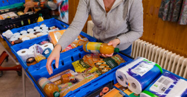 A person in a gray hooded sweatshirt reaches into a blue basket full of food at a food bank.