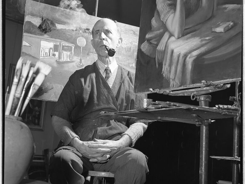 Black and white photo of an elderly man smoking a pipe in an art studio surrounded by paintings, brushes, and easels.