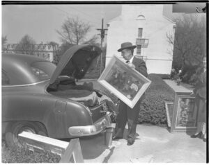 In this black and white photo, an elder man in a brimmed hat stacks paintings into the trunk of his car.