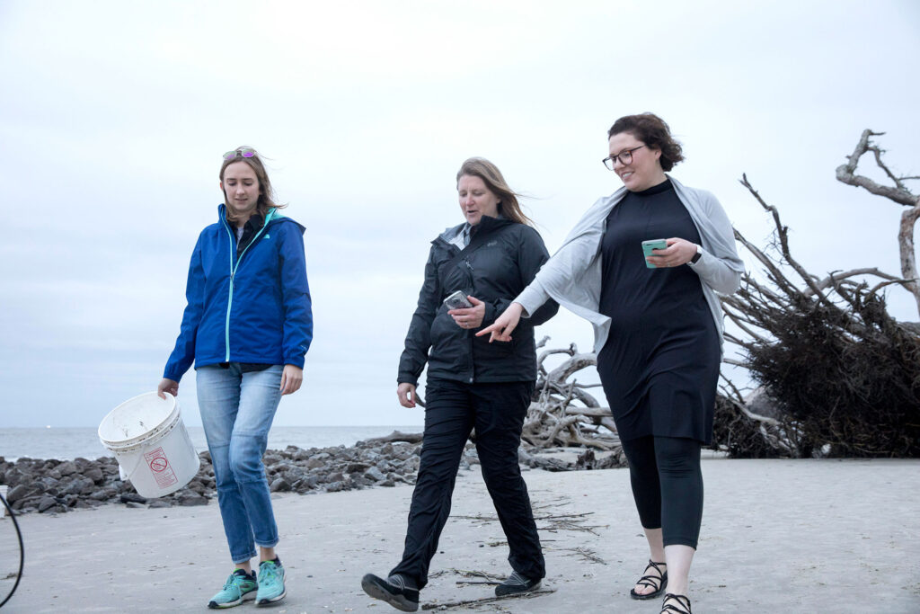 Three women walk across a beach collecting litter under overcast skies.