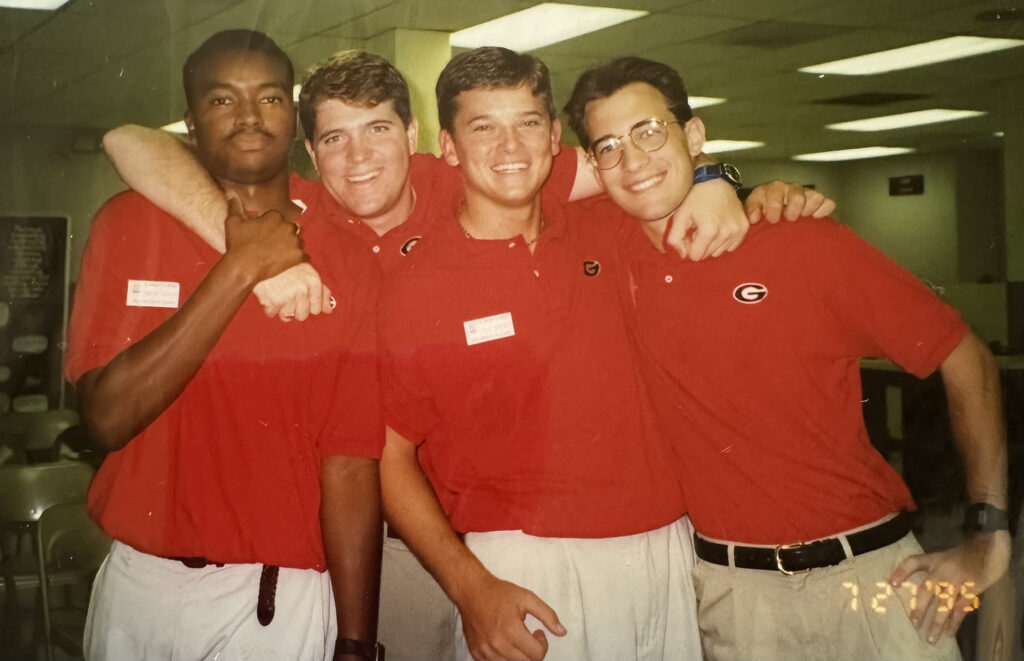 An older photo of four male orientation leaders at the University of Georgia. They are all wearing red shirts, white name tags, and light-colored pants.