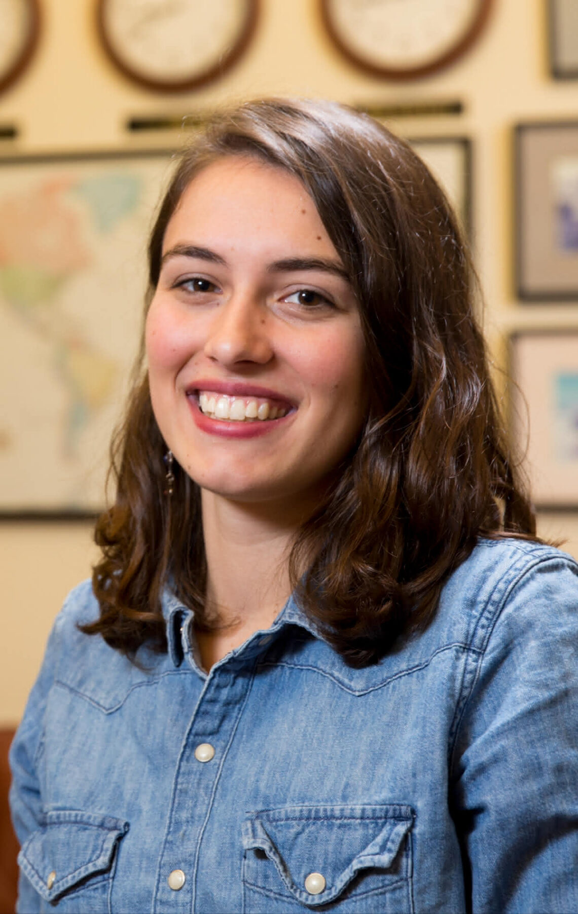 Portrait of UGA Rhodes scholar Laura Chourchesne, smiling in front of a wall of maps and clocks displaying times across the globe