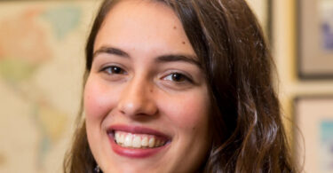 Portrait of UGA Rhodes scholar Laura Chourchesne, smiling in front of a wall of maps and clocks displaying times across the globe