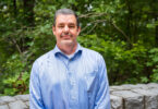 Environmental portrait of male researcher in a blue shirt in front of greenery.