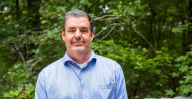 Environmental portrait of male researcher in a blue shirt in front of greenery.