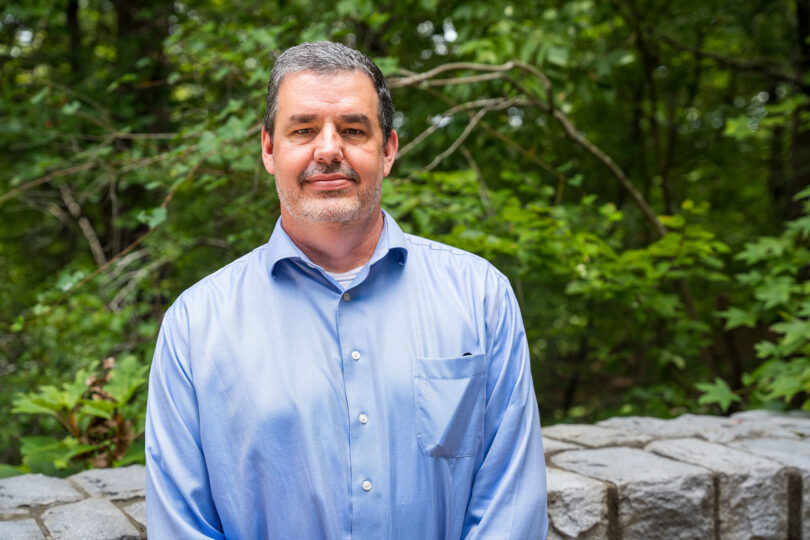 Environmental portrait of male researcher in a blue shirt in front of greenery.