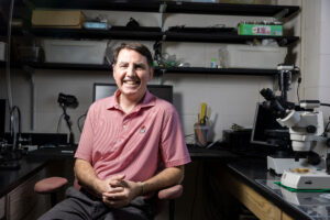 Photographed in his laboratory, a seated male researcher smiles at camera in a red and white striped polo shirt with a University of Georgia bulldog on it.