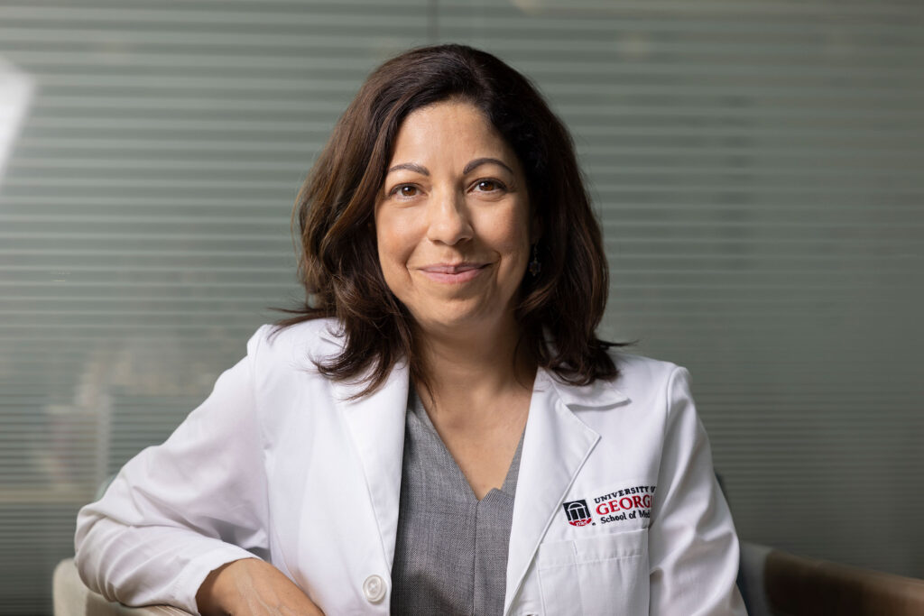 A female researcher in a UGA School of Medicine white lab coat smiles directly into the camera.