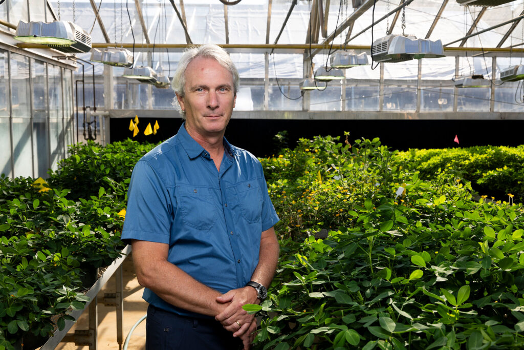 Environmental portrait of GRA Eminent Scholar, Scott Jackson, in a blue short-sleeved button down shirt in a greenhouse with rows of plants behind him.