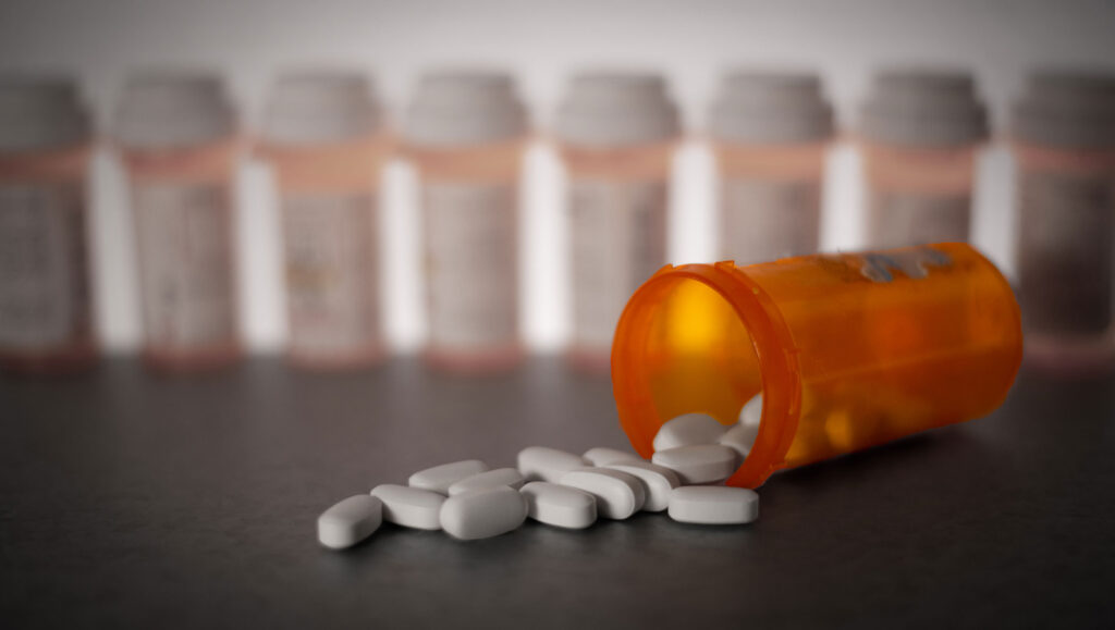 Pills are shown strewn across a tabletop spilling from a bottle with other prescription bottles lined up in the background.