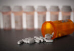 Pills are shown strewn across a tabletop spilling from a bottle with other prescription bottles lined up in the background.