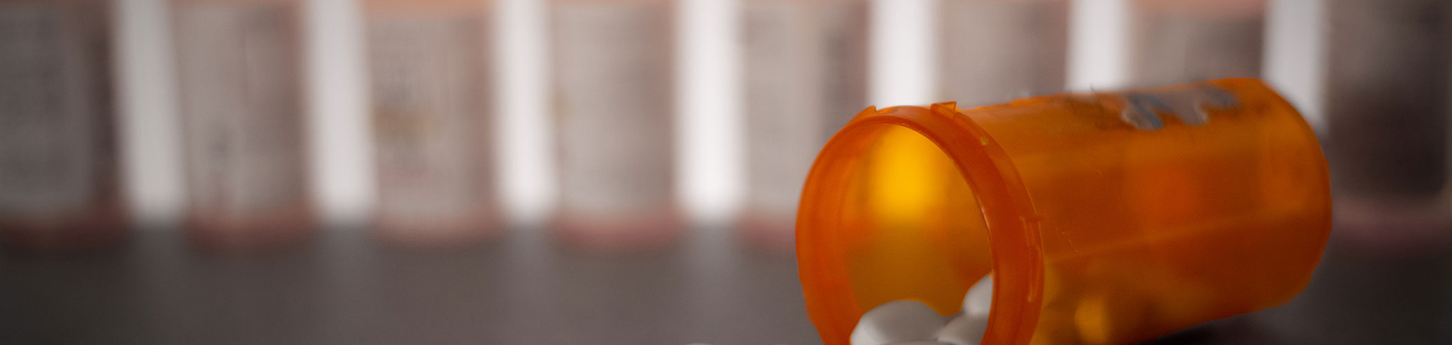 Pills are shown strewn across a tabletop spilling from a bottle with other prescription bottles lined up in the background.