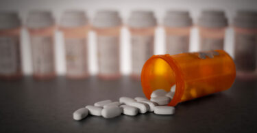 Pills are shown strewn across a tabletop spilling from a bottle with other prescription bottles lined up in the background.