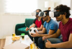 Three men in virtual reality headsets sit on a couch watching a virtual soccer game.