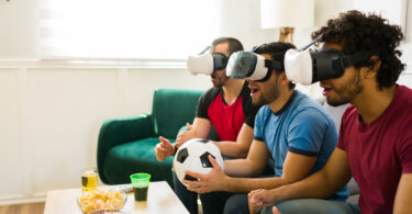 Three men in virtual reality headsets sit on a couch watching a virtual soccer game.