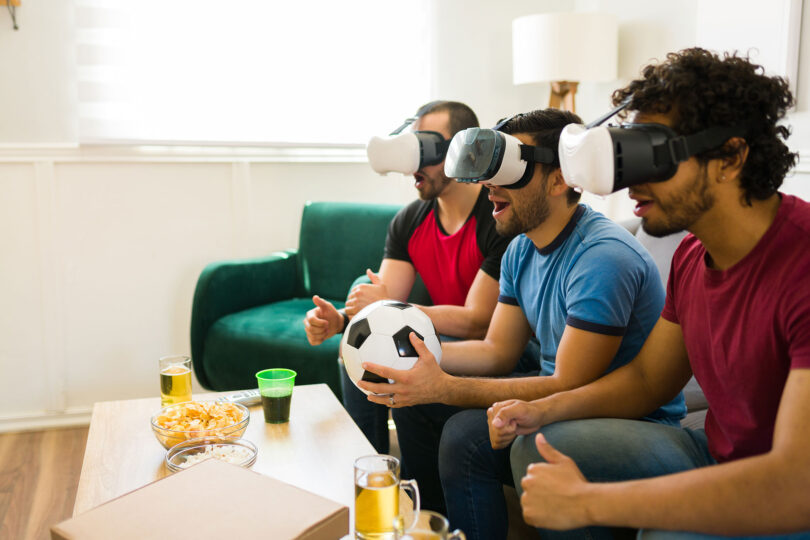 Three men in virtual reality headsets sit on a couch watching a virtual soccer game.