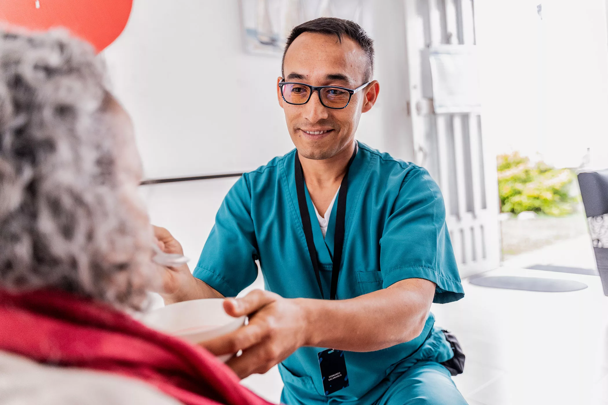 A male health care professional feeds an older woman a bowl of food.
