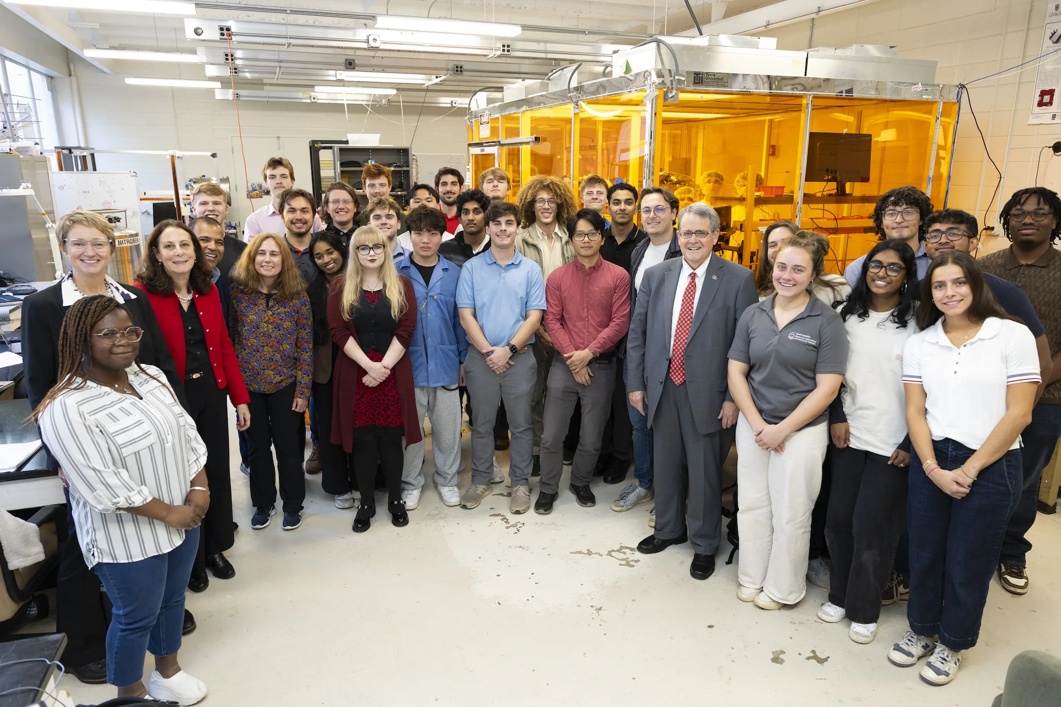 University of Georgia President Jere W. Morehead stands with students, faculty, and staff in the Small Satellite Research Laboratory during a tour celebrating the lab’s fifth anniversary, with research equipment and a cleanroom visible in the background.