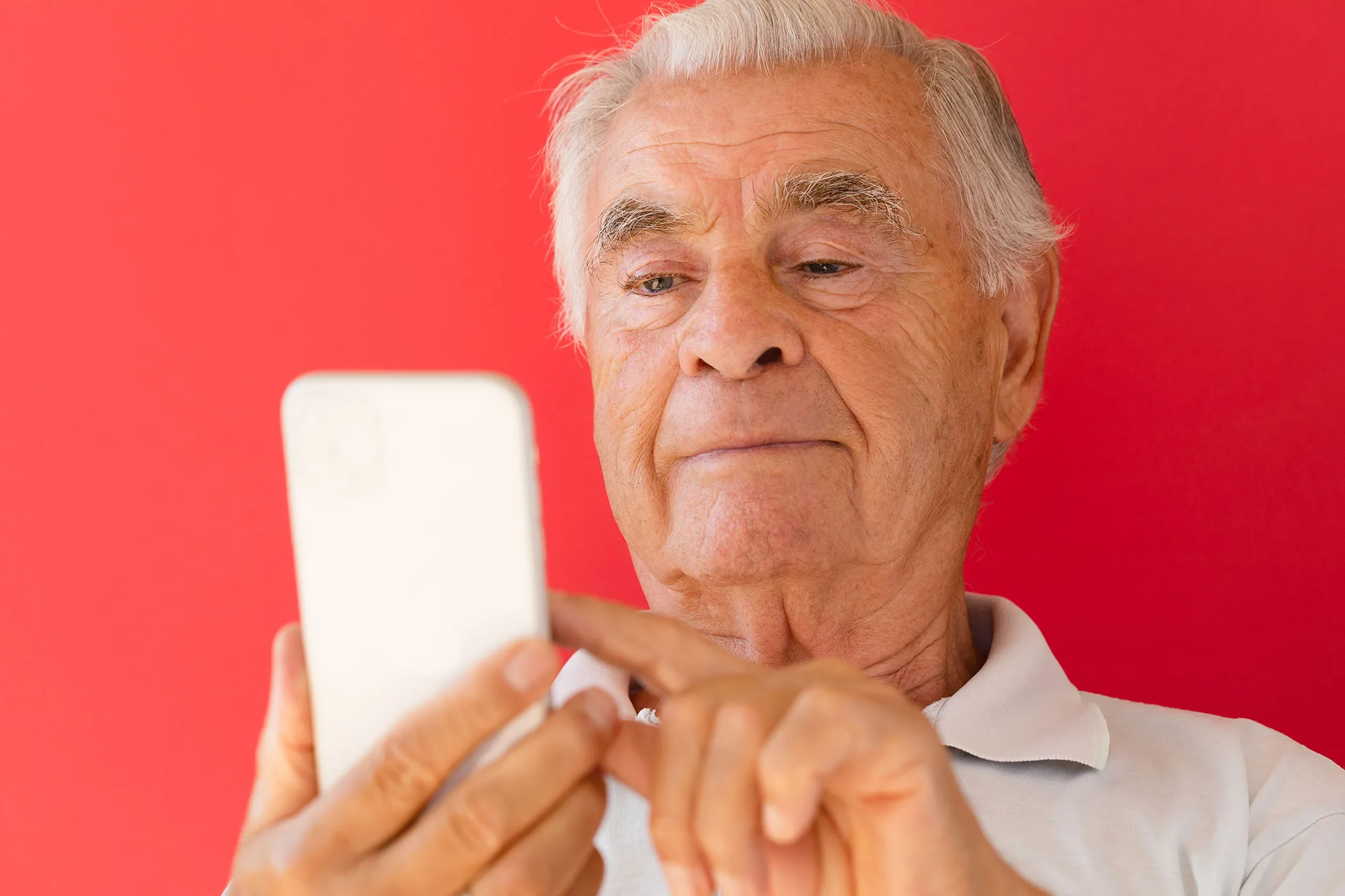 An older man types on a cell phone in front of a bright red background.