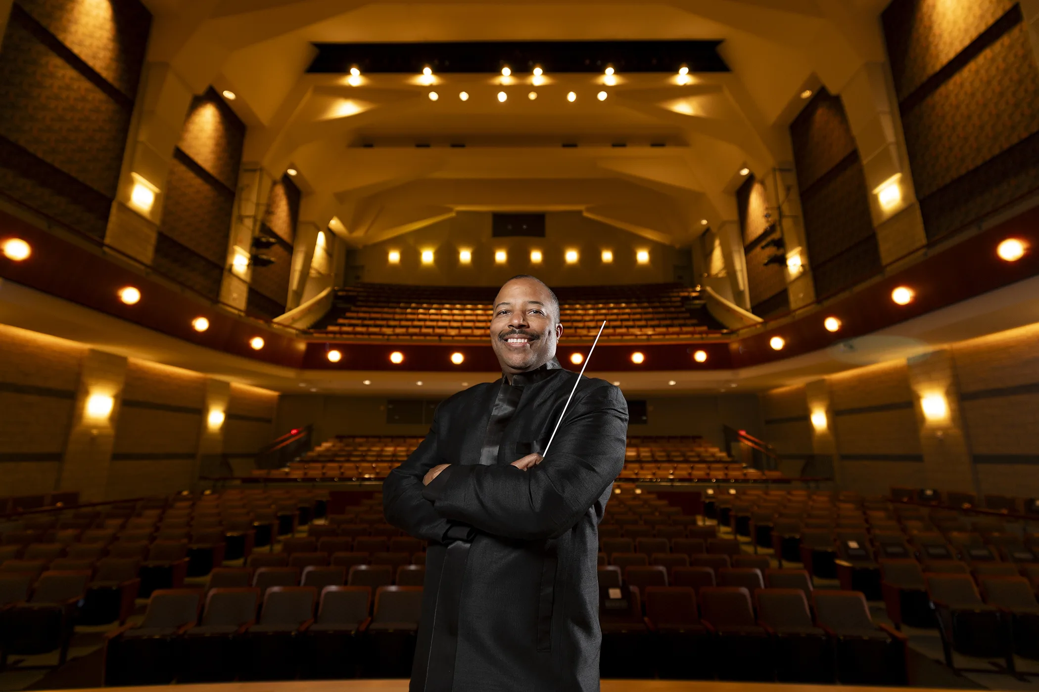 A conductor stands with his arms crossed, smiling at the camera. An empty concert hall opens behind him.