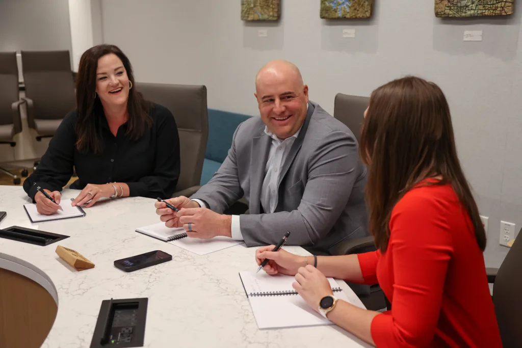 Three business professionals sit at a round conference table.