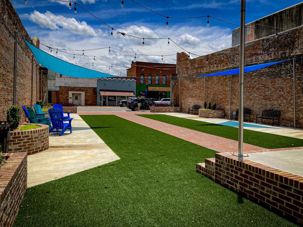Outdoor courtyard with artificial turf, brick walkways, string lights, and seating under blue shade sails.
