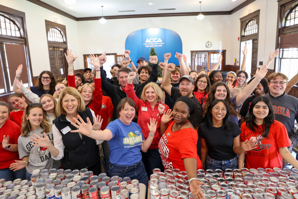 Volunteers gathered indoors around a table of canned goods, raising their hands for a group photo.