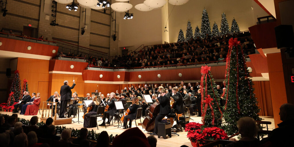 Orchestra members perform on a stage filled with holiday decorations.