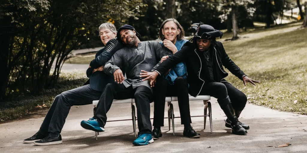 A group of four men pile together on three chairs along a walkway in a park.