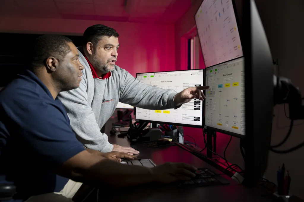 Two men look at a computer screen with the man on the right pointing to something on that screen.
