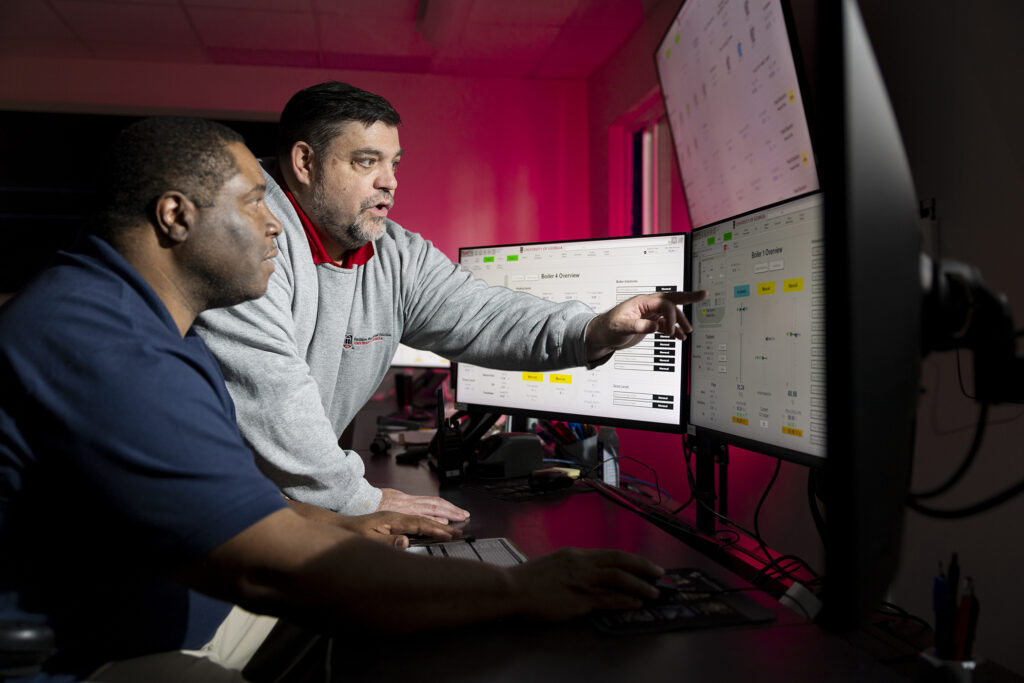 Two men look at a computer screen with the man on the right pointing to something on that screen.