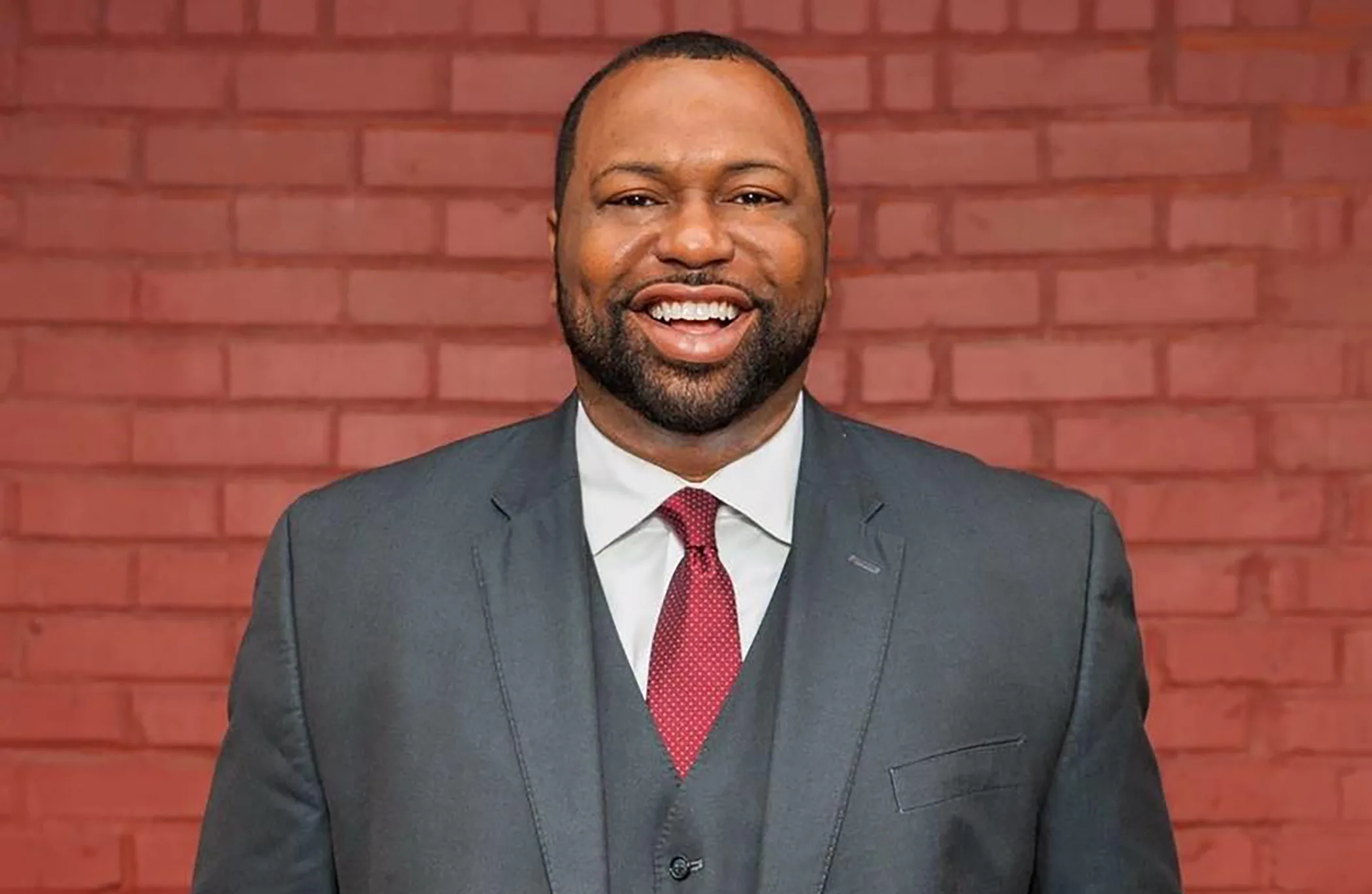 A man wearing a gray suit stands against a red brick wall.