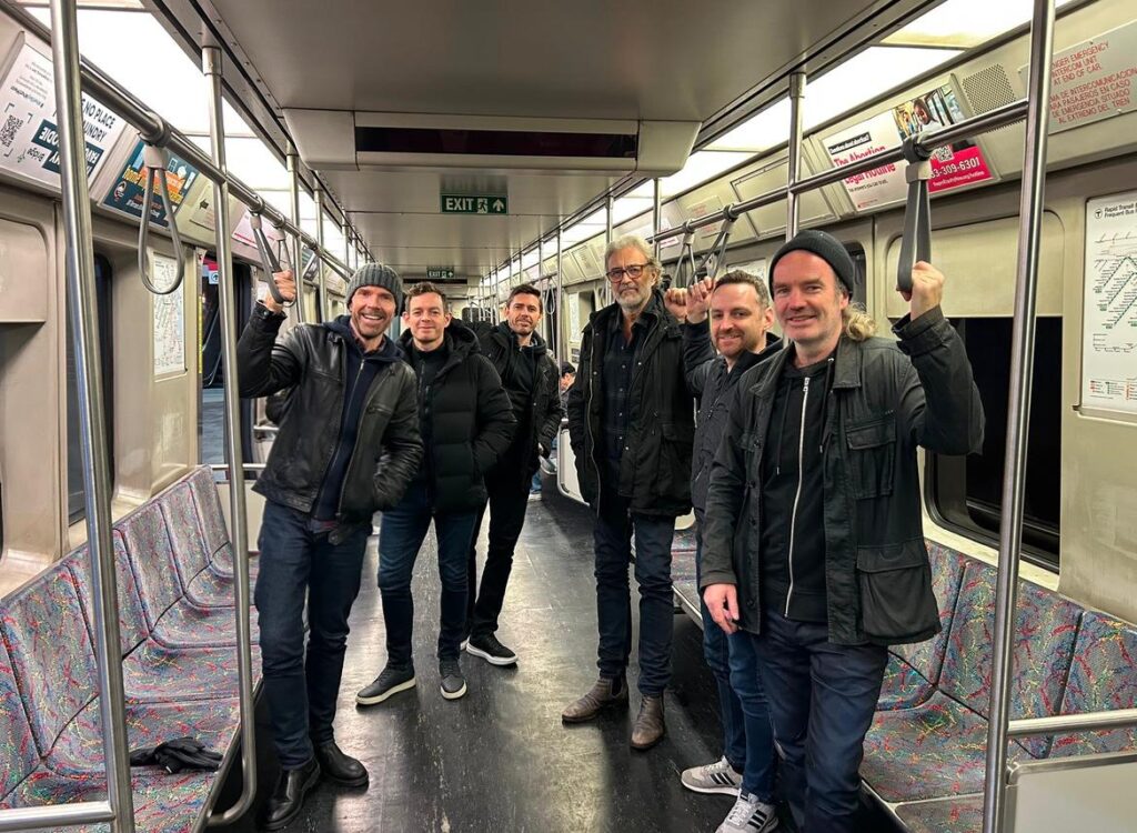 A group of six men, all wearing black jackets, stand in a subway car holding the upper grips.