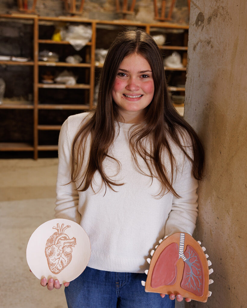 Student holds ceramic heart and lung pieces inside Cortona studio lined with shelving.