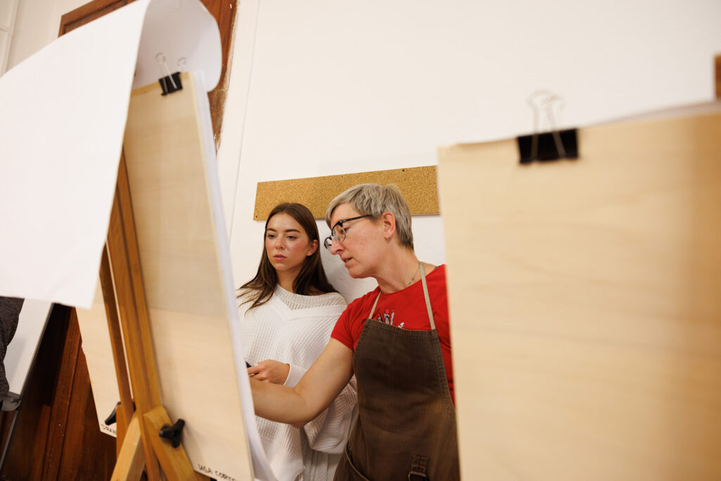 Instructor demonstrates drawing techniques beside student at easels in bright figure-drawing studio.