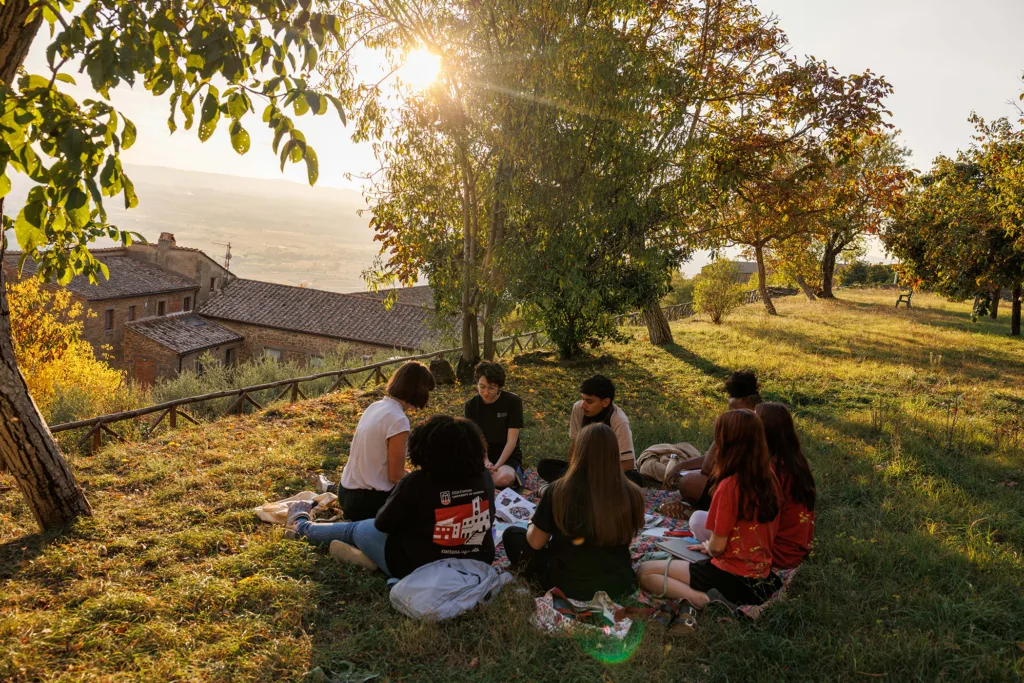 Students gather on hillside lawn under trees for outdoor class overlooking a valley.