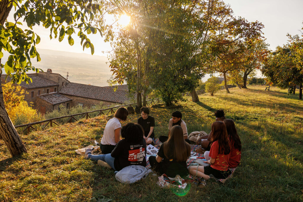 Students gather on hillside lawn under trees for outdoor class overlooking a valley.