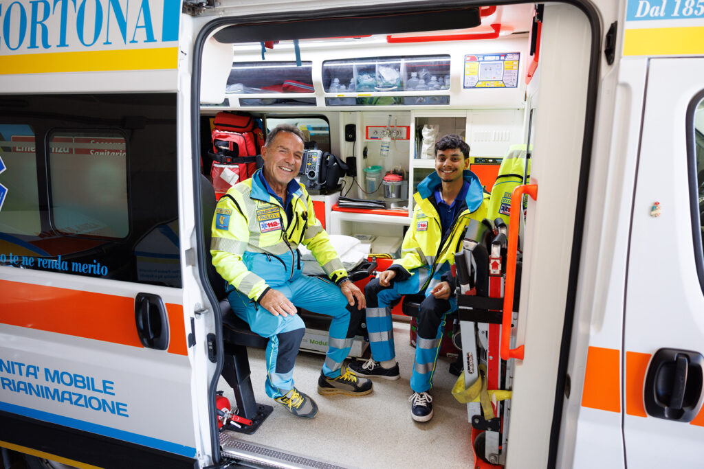 Student sits smiling with emergency staff inside brightly lit Cortona ambulance after shift.