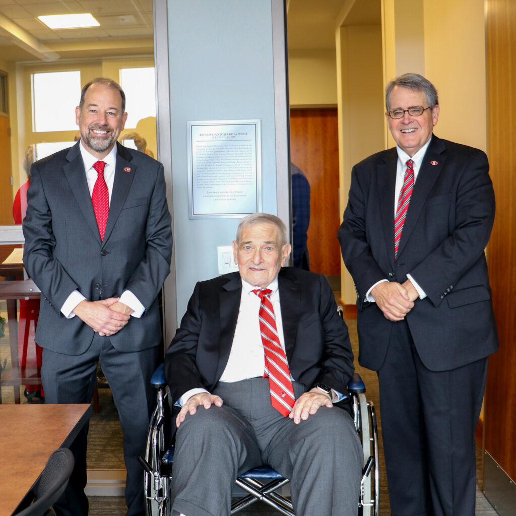 Two suited men stand beside older man in wheelchair in front of commemorative plaque.
