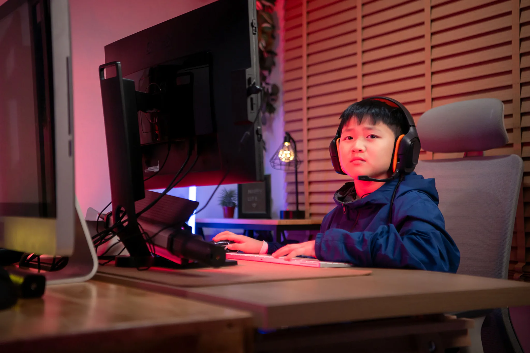 A boy gamer wearing headphones and playing on a desktop computer in a colorful gaming setup.