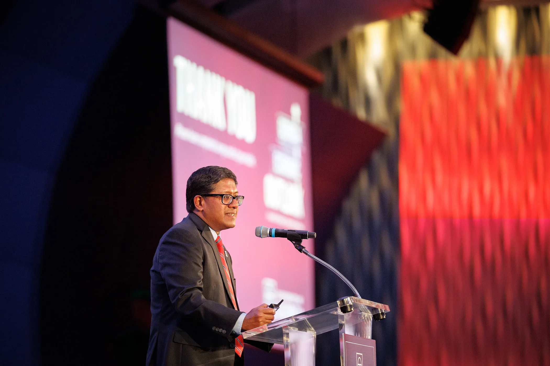 Speaker stands at clear podium with microphone, presenting before large illuminated event screens.