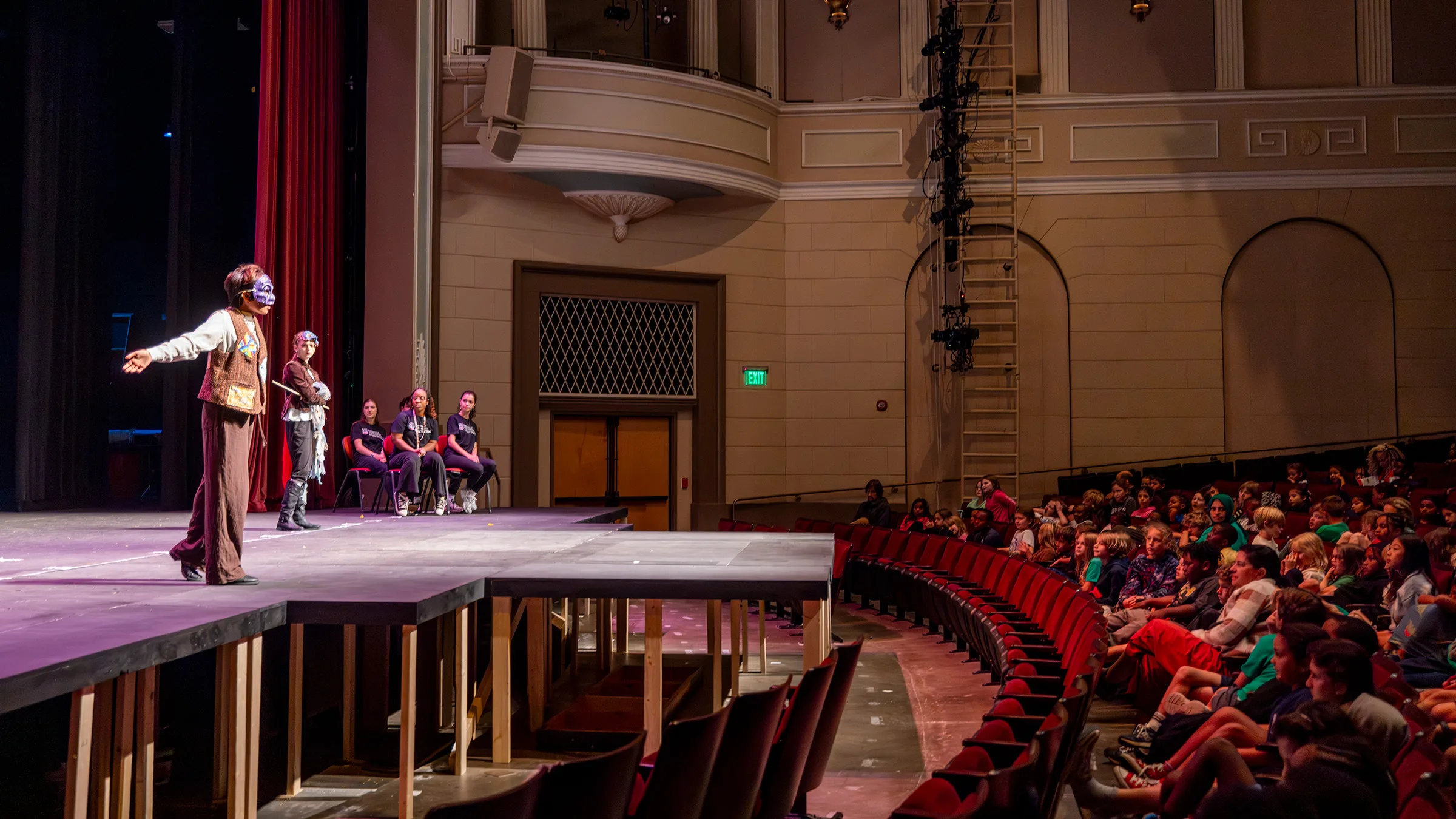 Student performers in masks act onstage as a large audience of children watches from red seats.