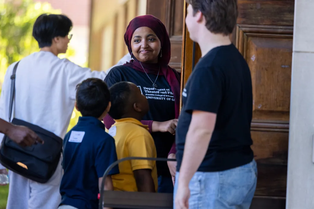 Student volunteers greet visiting children at theater entrance, smiling warmly as they arrive.