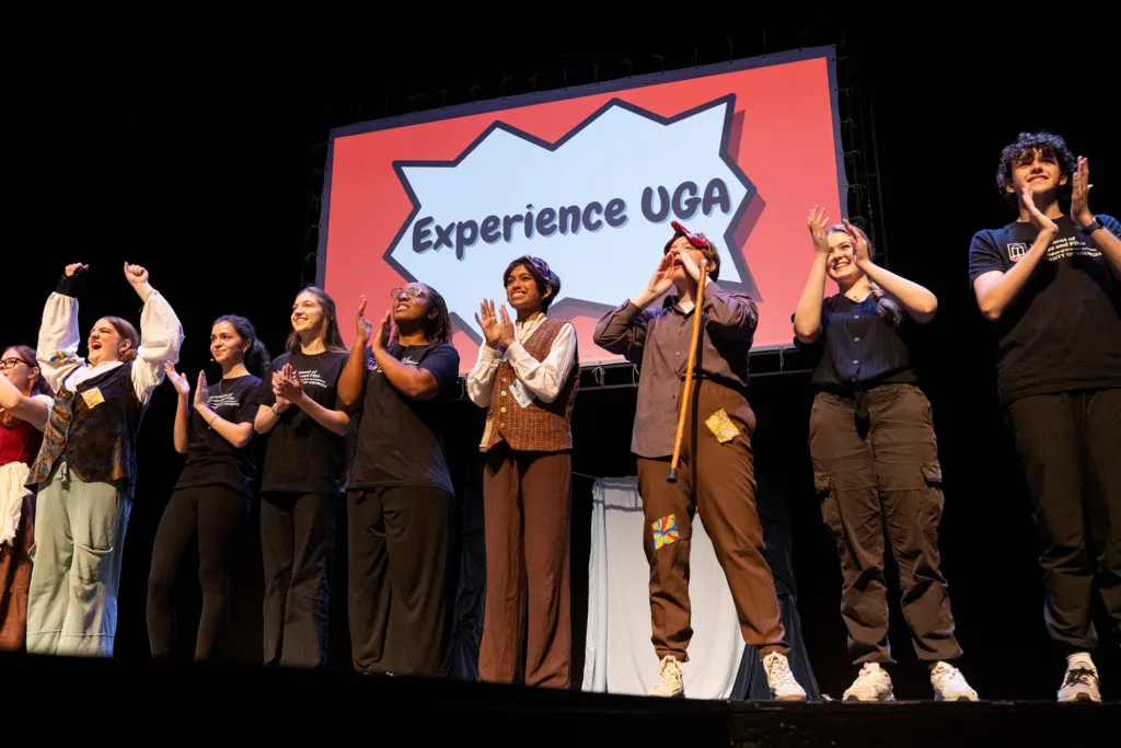 Student performers stand in a row onstage, cheering and clapping beneath a large “Experience UGA” sign.