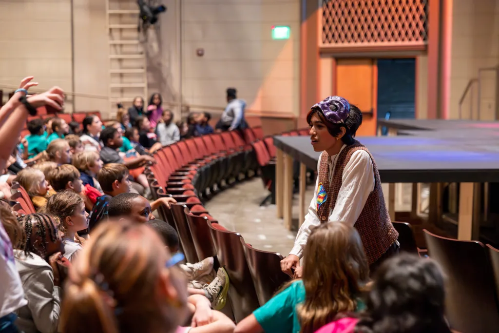 Student in colorful mask leans toward excited children in audience for post-performance conversation.