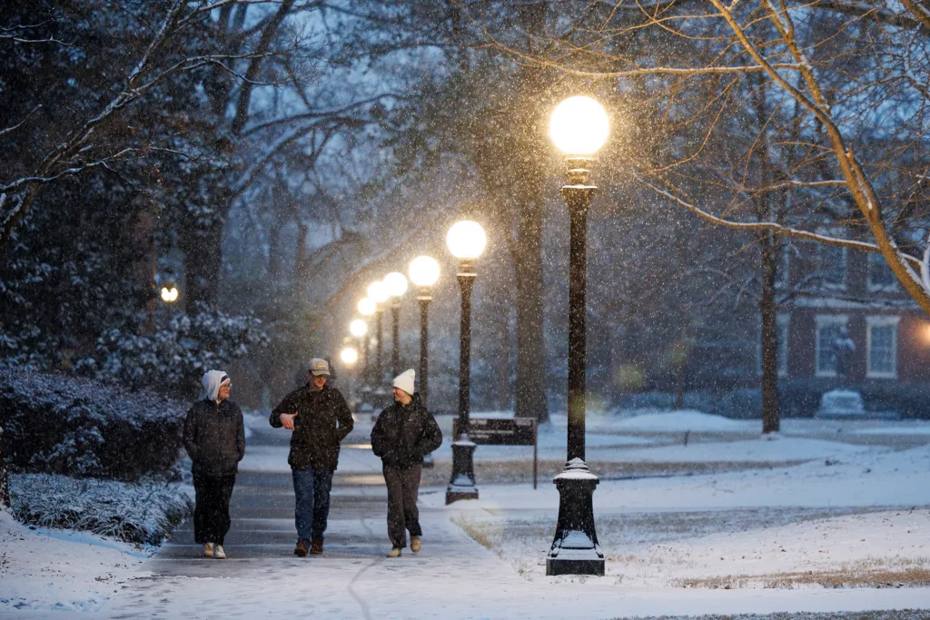 Students walk beneath glowing lampposts on snowy North Campus path during peaceful evening snowfall.