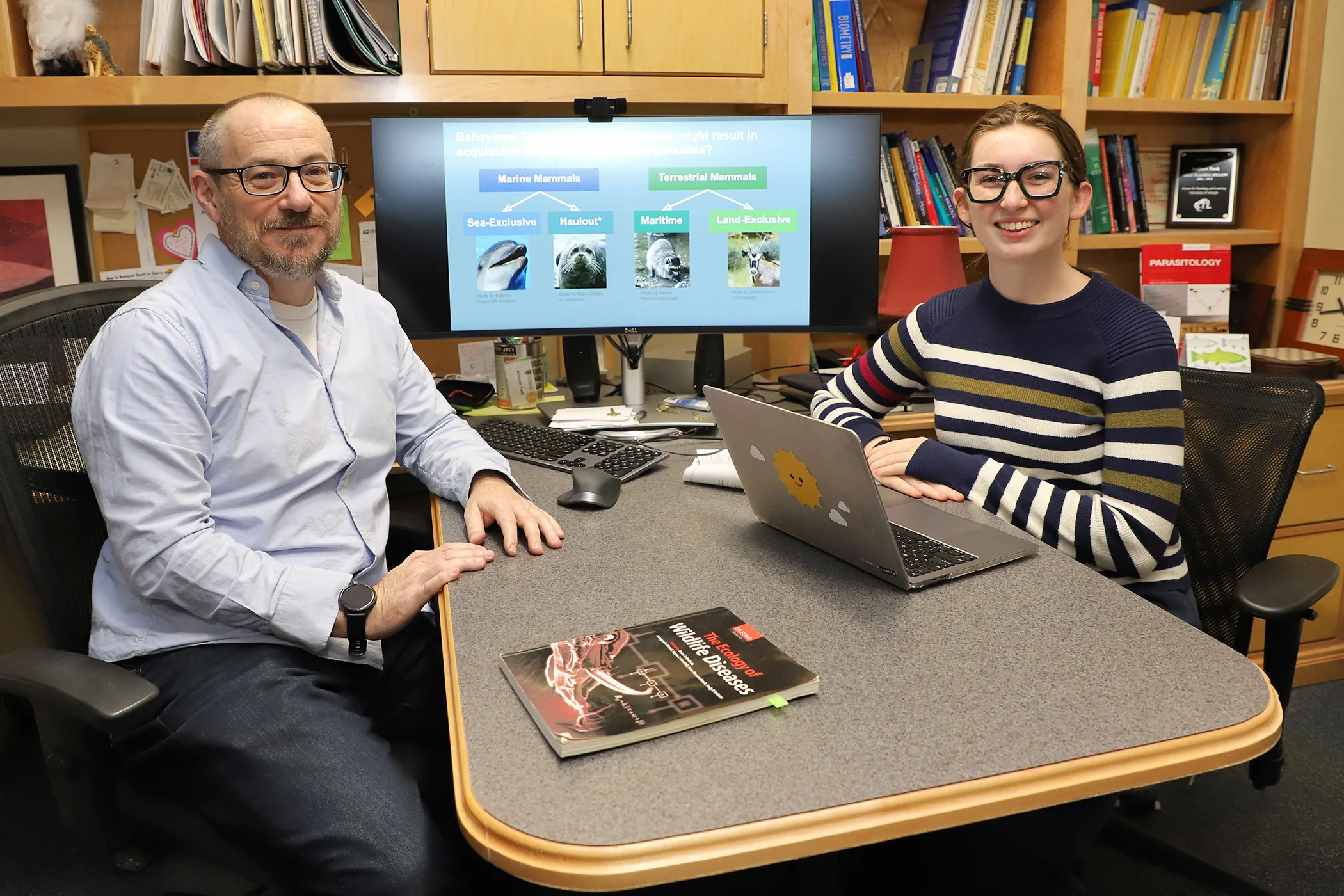 Student in striped sweater with laptop meets with faculty mentor in office.