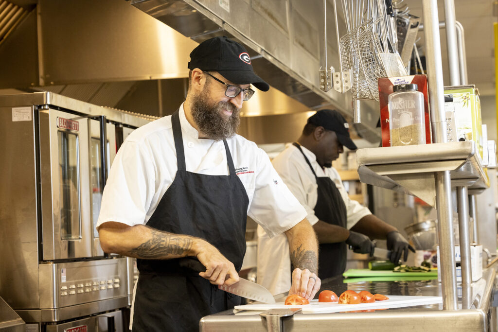 A man in a UGA Dining Services chef's outfit stands in an industrial kitchen, slicing tomatoes. 