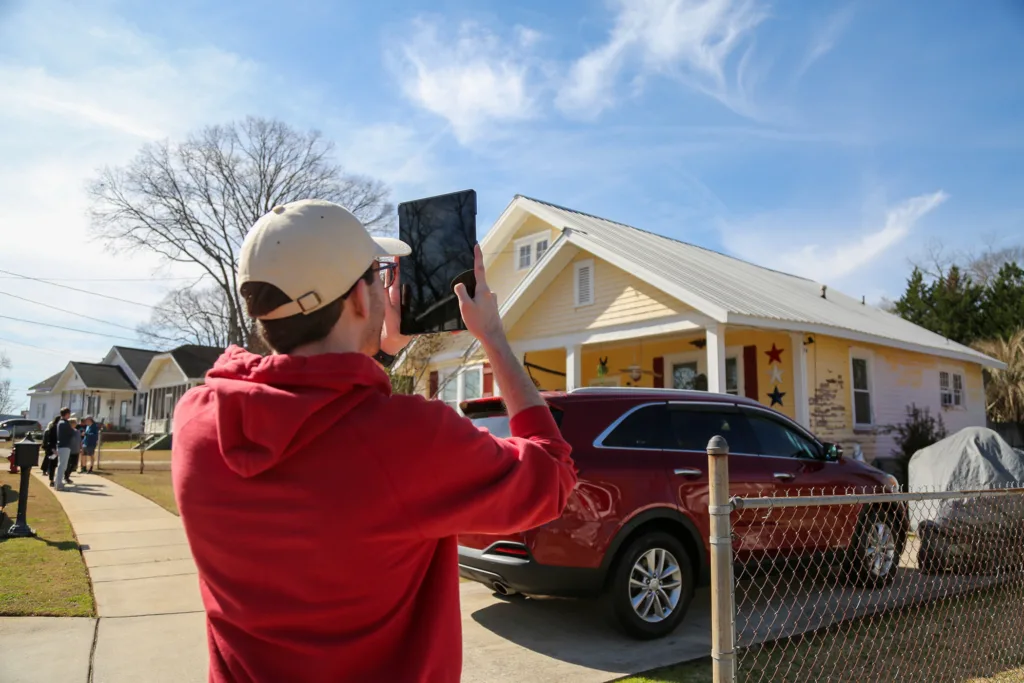 A student photographs a yellow house while surveying a neighborhood during a community housing assessment.