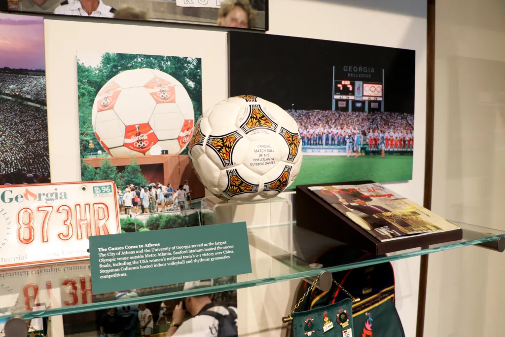 Official match ball from the 1996 Atlanta Olympic Games displayed alongside photographs and memorabilia highlighting soccer competitions hosted in Athens at University of Georgia venues.